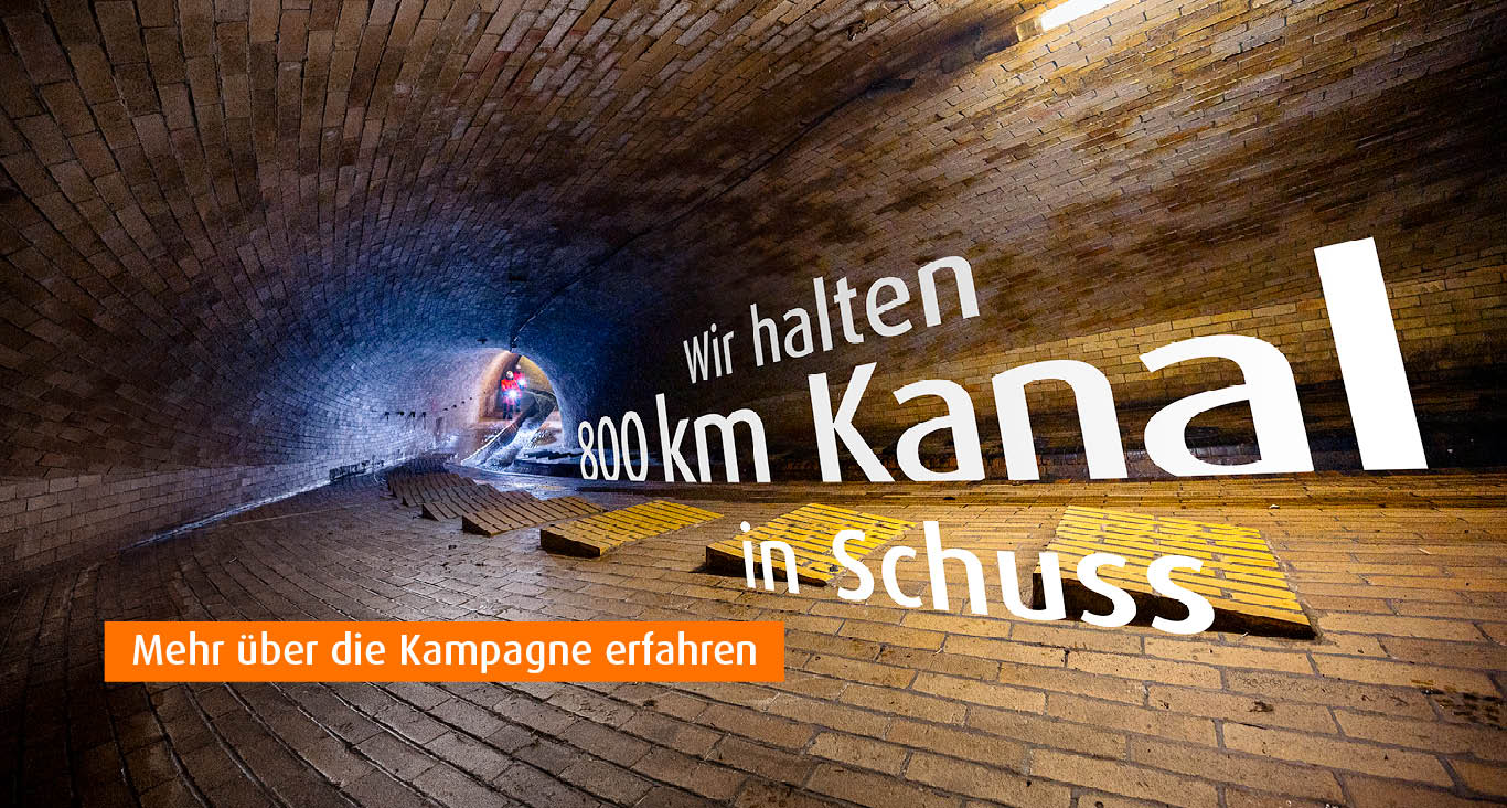 Blick in einen gemauerten Abwasserkanal mit zwei Personen in oranger Schutzkleidung mit Helmen und Taschenlampen im Hintergrund. Text auf dem Bild: Wir halten 800 km Kanal in Schuss. Mehr über die Kampagne erfahren.
