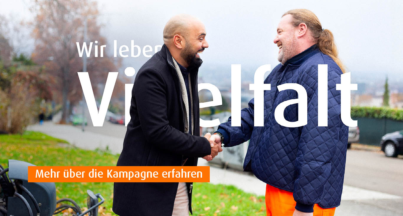 Zwei Männer begrüßen sich per Handschlag auf einer Straße, einer kahlköpfig, einer mit Bart und langen Haaren, beide lächeln. Text auf dem Bild: Wir leben Vielfalt. Mehr über die Kampagne erfahren.